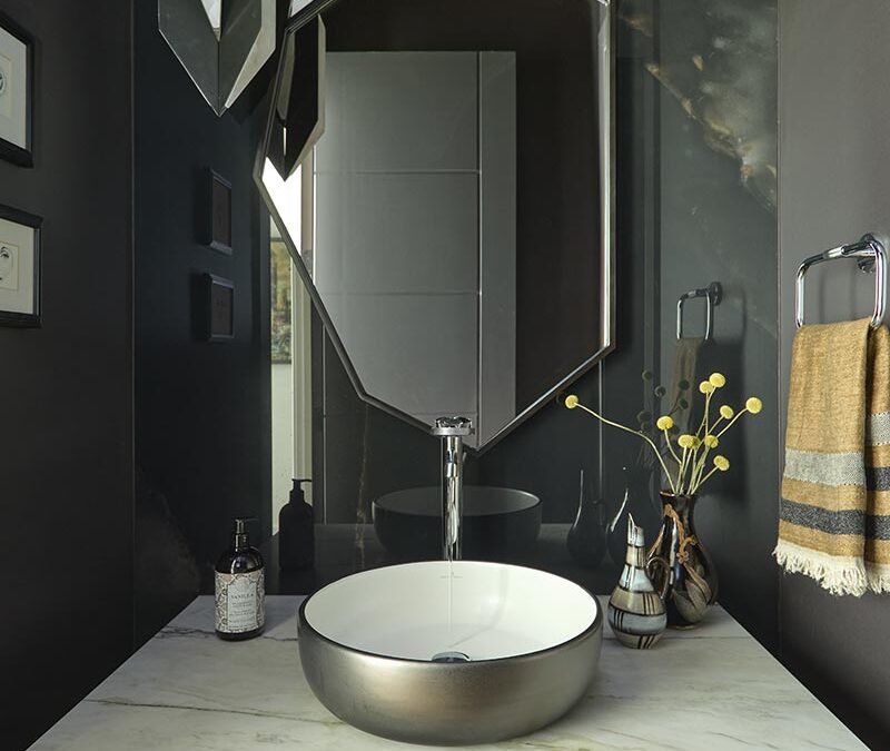 Modern bathroom vanity with a round vessel sink, geometric wood cabinet, octagonal mirror, pendant light, and dark walls with marble accents. Towel hangs on the right, soap and decor on the counter.