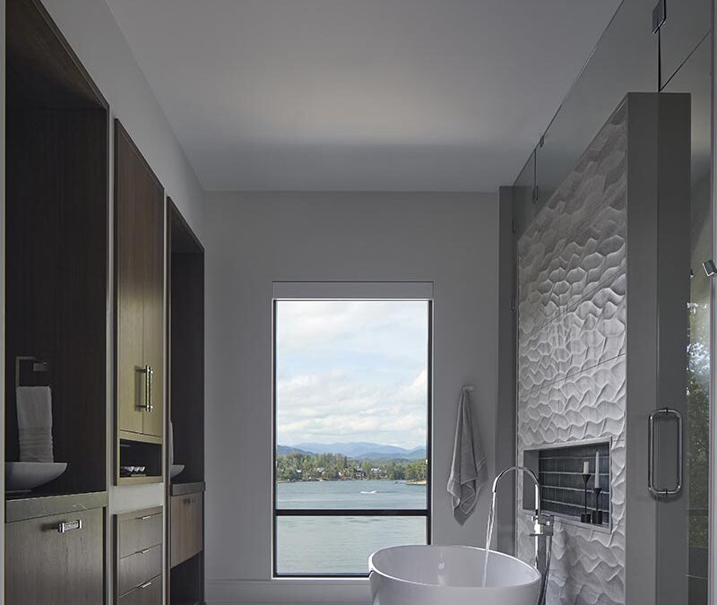 Modern bathroom with a freestanding white bathtub, textured wall, built-in shelves, and a large window overlooking lake keowee and mountains.